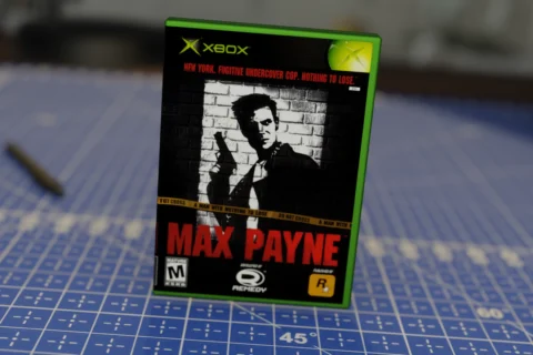 Carátula del videojuego Max Payne para Xbox, con un diseño en blanco y negro de un hombre armado de espaldas a una pared de ladrillos, con su nombre en rojo y una cinta de precaución cruzando la portada.