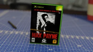 Caja del videojuego Max Payne para Xbox en primer plano, con un fondo desenfocado que muestra una superficie cuadriculada y una herramienta.