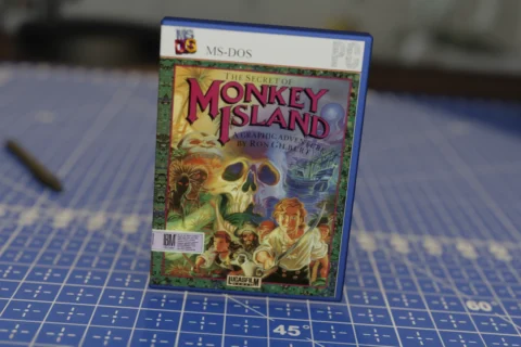 Caja del videojuego The Secret of Monkey Island para MS-DOS en una funda de plástico azul, colocada sobre una superficie azul cuadriculada con herramientas desenfocadas en el fondo.